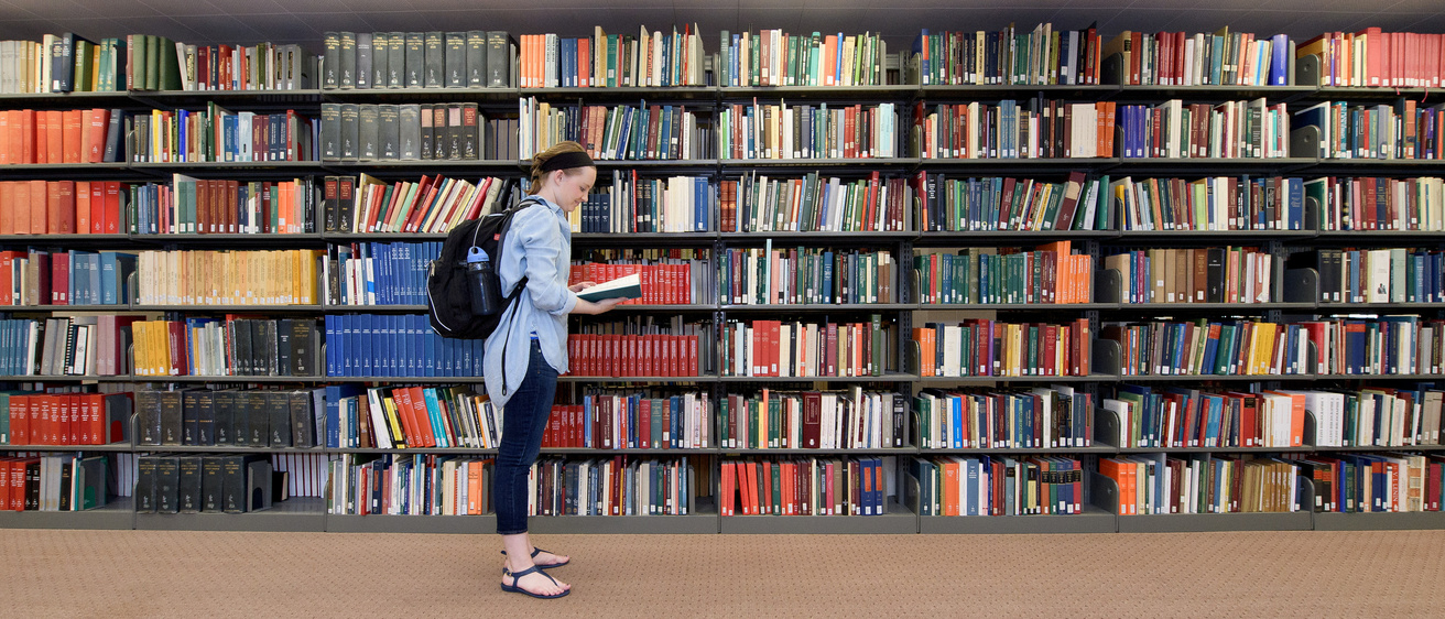 A woman stands in front of a long shelf full of books while looking through a book